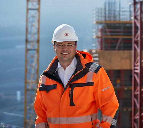 Bauingenieur Deutsche Bahn Bauingenieur Jörg steht mit orangener Sicherheitsweste und Helm vor seiner Baustelle. Im Hintergrund steht ein großer Kran.